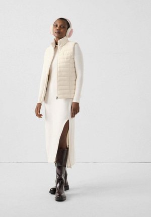 Beige gewatteerde gilet over een roomkleurige jurk met een zijventilatie, gecombineerd met bruine kniehoog laarzen. Lichtroze oorkappen maken de outfit compleet.