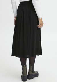 Sort midi-nederdel med en samlet elastisk talje, parret med sorte tights og kraftige ankelstøvler med hæl. Glat stof med diskret drapering.