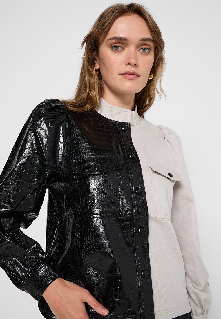 Koszula z kołnierzykiem, z jednej strony pokryta czarną, sztuczną teksturą krokodyla, a z drugiej gładkim, jasnoszarym materiałem. Wyposażona w zapięcia na guziki i kieszenie.