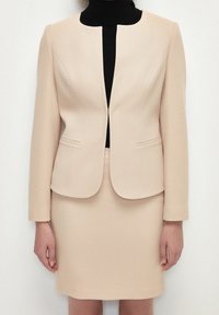 Beige räätälöity takki, jossa on pyöreä kaulus, kaksi etutaskua ja yhteensopiva hame, käytetty mustan poolopaidan päällä. Rakennekaava tekstuurilla.