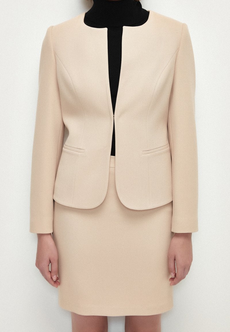 Beige räätälöity takki, jossa on pyöreä kaulus, kaksi etutaskua ja yhteensopiva hame, käytetty mustan poolopaidan päällä. Rakennekaava tekstuurilla.