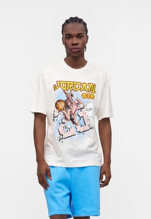 Jeune homme aux cheveux tressés portant un t-shirt blanc Jordan Air avec un graphisme représentant un joueur de basketball et un short bleu clair, debout devant un fond uni.