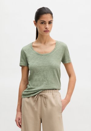 Kobieta z ciemnymi włosami związanymi z tyłu, ubrana w jasnozieloną koszulkę z krótkim rękawem i beżowe spodnie z sznurkiem, stojąca z jedną ręką w kieszeni.