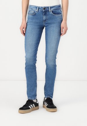 Tanke modre denim hlače z zavihanimi žepi in detajli šivanja. Obute so z črnimi supergah, ki imajo bele črte in svetlo rjav podplat.