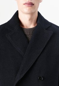 Musta kammattu villakangastakki, jossa on kaksoisnapitus ja leveät kaulukset, kerroksittain ruskean teksturoidun neulepaidann yli.