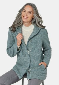 Tauchblaues Wollmischjackett mit Reißverschluss, Druckknöpfen und großem Kragen. Verfügt über strukturierten Stoff und Seitentaschen, getragen über einem cremefarbenen Pullover.