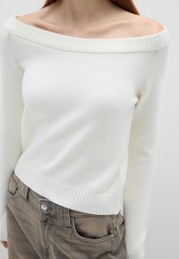 Weißer Off-Shoulder-Pullover mit geripptem Ausschnitt und Saum, aus weichem Material; kombiniert mit beigen, hochgeschnittenen Hosen mit einem Frontknopf.