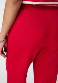 Pantalons élastiques rouges à taille haute avec detail de poche arrière. Texture lisse, forme fuselée et couleur unie sans motifs ni accents en métal.