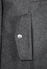 Gråt uldstof med en lomme, der har en metaltryklåsdetalje. Teksturen har en blød, børstet finish.