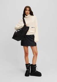 Weißer gerippter Rollkragenpullover, schwarzer Jeans-Minirock, schwarze klobige Schneestiefel mit Schnürsenkeln und eine große schwarze Tragetasche mit weißem Text.