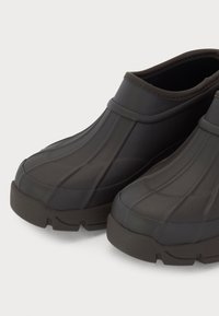 Pantofi din cauciuc negri, cu un design nervurat, vârf rotunjit și talpă groasă, texturată. Au o deschidere largă pentru a fi ușor de purtat.