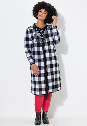 Manteau long à carreaux noir et blanc avec des franges, associé à une chemise noire à pois, un legging rouge et des bottes noires cloutées.