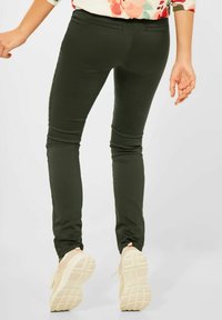 Pantaloni slim-fit di colore verde oliva con una texture liscia, dotati di due tasche posteriori e abbinati a sneakers chiare.