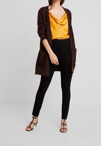 Hnědý chlupatý kardigan s otevřeným pletením, žlutý saténový top s krčním výstřihem, černé přiléhavé kalhoty a černé sandály na podpatku. Jednoduchý design s kapsami na kardiganu.