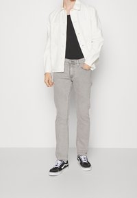 Veste en denim blanc sur une chemise noire, associée à un jean gris clair et des baskets noires. La tenue présente un style décontracté et ajusté.