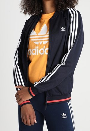 Personne portant une veste et un pantalon Adidas bleu marine avec des bandes blanches et un t-shirt Adidas orange, les bras croisés devant.