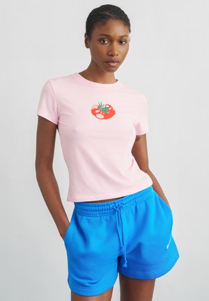 Kobieta ubrana w jasnoróżowy t-shirt z grafiką pomidora i jasnoniebieskie spodenki Nike, stoi z rękami w kieszeniach na jednolitym tle.