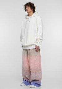 Felpa crema con tasca a marsupio e logo nero, abbinata a pantaloni larghi con gradiente pastello nei toni di rosa, blu e bianco. Scarpe da ginnastica bianche.