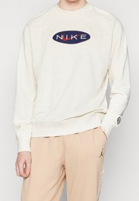 Krémová mikina s navy oválním logem, na kterém je bílé písmo "NIKE". Dlouhé rukávy a spodní lem jsou žebrované, mikina je vyrobena z jemné látky.