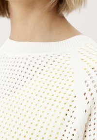 Biały dziany sweter z wyraźnym wzorem z małymi otworami i żółtymi akcentami, podkreślający jego przewiewną fakturę i design.