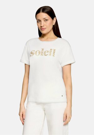 Kvinde med glat brunt hår bærer en hvid T-shirt med ""soleil"" i gyldne bogstaver og matchende hvide bukser, stående mod en ensfarvet baggrund.