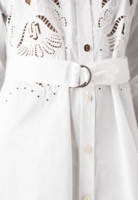 Camicia bianca con ricami dettagliati e fori. Presenta una cintura coordinata in tessuto con un anello in metallo. Design con bottoni a vista.