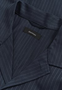 Mornarsko modra tkanina z belimi tankimi črtami, ki prikazuje zložen ovratnik in črno etiketo z napisom "Matinique." Gladek otip, krojen dizajn.