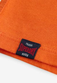 Oranžová tkanina s textúrovaným povrchom, s tmavomodrou štítkou s nápisom "SUPERDRY" v červenej a bielej farbe, umiestnenou na leme.