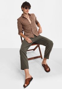 Camicia a righe marrone con bottoni, pantaloni oliva e sandali marroni con cinturini incrociati. Modello seduto su una sedia di legno contro uno sfondo neutro.