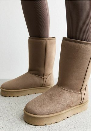 Bottes mi-mollet en daim doux couleur noisette avec semelles plates en caoutchouc, portées avec des leggings bruns côtelés sur un fond de sol clair.