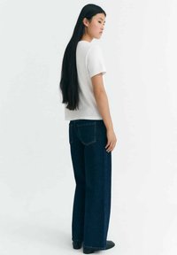 Weißes T-Shirt mit kurzen Ärmeln, kombiniert mit dunkelblauen weiten Jeans. Langes schwarzes Haar fällt glatt den Rücken hinunter. Minimalistisches Design und Passform.