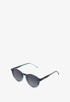 Des lunettes de soleil rondes teintées de bleu avec des verres à dégradé sombre et des branches fines, arborant le logo "KOMONO" sur le côté.