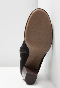 Bottine noire en daim avec une semelle en caoutchouc marron, dotée d'une fermeture éclair sur le côté et d'un talon empilé. La texture est lisse avec une finition mate.