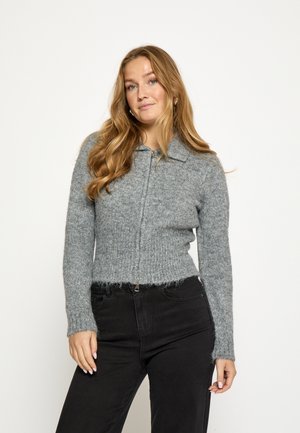 Grijze, geribbelde gebreide trui met een ritssluiting aan de voorkant en een kraag. Heeft een zachte textuur en een rafelige zoom. Gecombineerd met een zwarte jeans.