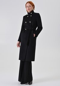 Zwarte wollen jas met een hoge kraag, dubbelknopen met gouden knopen, zijzakken en een tailleband, gecombineerd met zwarte wijde pantalon.