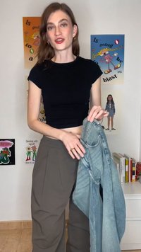 Jauna sieviete melnā īsā topā un brūnās biksēs, turot rokās džinsa jaku, stāv telpā ar krāsainiem plakātiem un grāmatām.