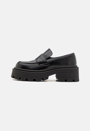 Mocassins noirs en similicuir avec une semelle plateforme épaisse et texturée, et une large bride sur le dessus pour un maintien sûr et un style assuré.