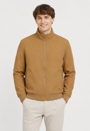 Lächelnder Mann in einer beigefarbenen Zip-Jacke mit Taschen und hellbeigen Hosen steht mit den Händen in den Taschen vor einem schlichten Hintergrund.