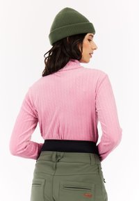 Lange mouw roze trui met kabelpatroon, hoge kraag; gecombineerd met olijfgroene broek met een zwarte tailleband en zakken.