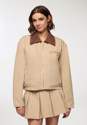 Giacca corta beige con colletto in velluto a coste marrone, zip frontale e una piccola tasca sul petto con zip. Tessuto morbido, vestibilità comoda.
