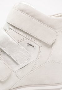 Zbliżenie na beżowy zamszowy i metaliczny skórzany sneaker z dwoma paskami na rzep i małym logo GEOX na bocznej części.