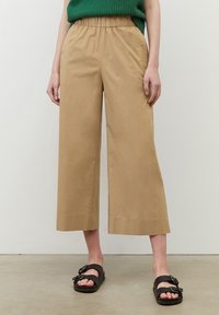 Beige beskurna vida byxor med elastisk midja och sidofickor, tillsammans med svarta sandaler med spännband.