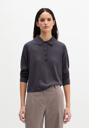 Vrouw met lang donker haar, gekleed in een donkergrijs poloshirt met lange mouwen en beige hoge taillebroek, staand tegen een witte achtergrond.