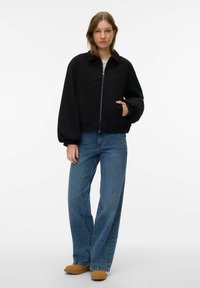 Veste bomber noire raccourcie avec fermeture éclair à l'avant, dotée d'un col, de poignets élastiques et de poches latérales, portée avec un jean bleu à jambes larges et des chaussures marron.