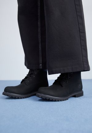 Bottines noires à lacets portées avec un pantalon noir à jambes droites sur un sol bleu clair avec un fond uni.