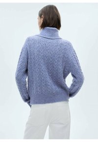 Jersey de punto lila con cuello alto y ancho y patrón texturizado de trenzas. Presenta un ajuste relajado y bajo acanalado, combinado con pantalones blancos.