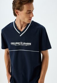 Marineblå bomulds T-shirt med V-hals, med hvide striber ved udskæringen og et "College League" grafik i hvid på brystet.