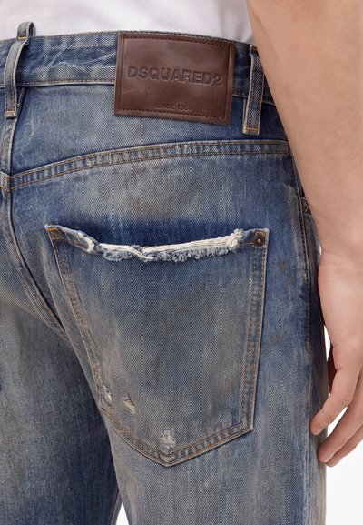 Gros plan sur un jean bleu usé avec une poche arrière effilochée et un patch en cuir marron DSQUARED2 au-dessus de la poche.