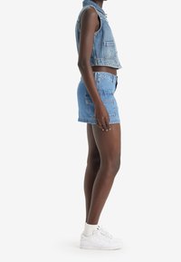 Denimväst och shorts i ljusblått. Västen har två framfickor; shortsen har en fransad nederkant och bakficka. Vita sneakers med synliga strumpor.