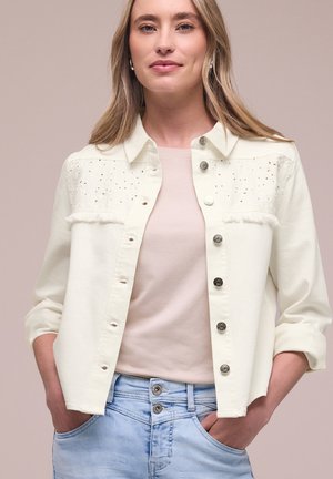 Mujer con chaqueta bordada color beige claro abotonada sobre una camiseta beige con vaqueros azul claro con doble botón, manos en los bolsillos.